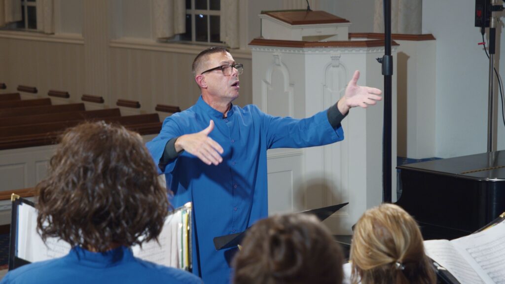 Photo of Christopher, in blue choir robe, conducting a church choir